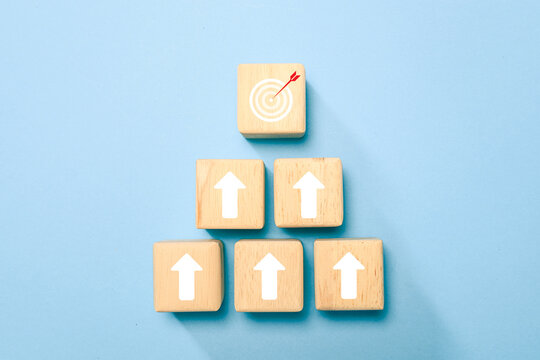 Wooden blocks with upward arrows lead to a target and arrow, symbolizing goal achievement, growth, strategy, success and business progress on a blue background with ample copy space.