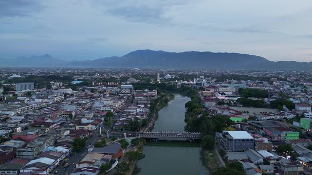 Banda Aceh, Indonesia : Orbit aerial drone footage of riverside mosque and residential area in Banda Aceh of Sumatera, Indonesia toward Baiturrahman Grand Mosque and mountain under cloudy sunset sky