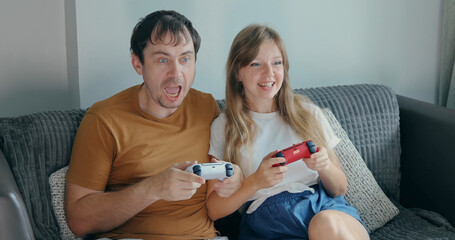 Man and woman playing games on grey sofa, holding gamepads with excited expressions, indoor home entertainment lifestyle, couple leisure activity at home. © TravelMedia