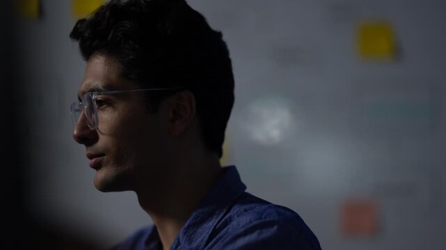 A young engineer in profile stands against the flowchart wall and listens without interrupting inside a dimly lit artificial intelligence startup office at night.
