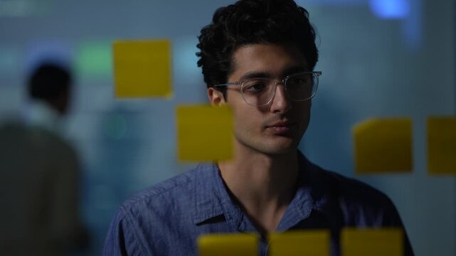 A young engineer behind a note covered pane reads the plan through the glass while a colleague works the notes beside him inside a dimly lit machine learning startup office.