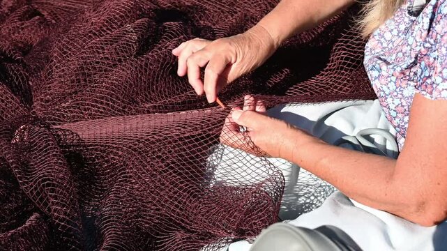 A woman sitting on a boat and preparing a fishing net, with different pieces of netting and equipment visible around her. Bright natural sunlight emphasizes the intricate mesh texture and details of t