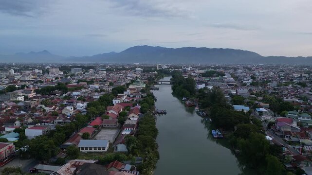 Banda Aceh, Indonesia : Forward aerial drone footage of riverside mosque and residential area in Banda Aceh of Sumatera, Indonesia toward Baiturrahman Grand Mosque and mountain under cloudy sunset sky