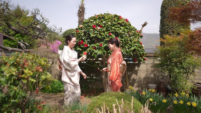 Two people in patterned kimono look toward a red ball hanging from a handheld string toy in a garden.