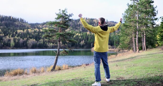 A person stands on a grassy bank, arms raised in joy, surrounded by stunning trees and a serene lake. The sun shines brightly, highlighting the beauty of the moment.