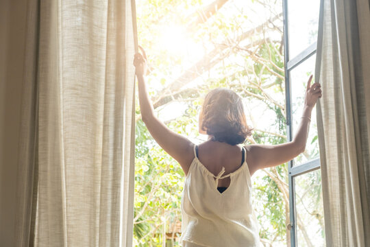 The woman was at the window in the bedroom. She opened the curtains on the window. In the morning and she looks at the view of mountains and trees at sunrise.	