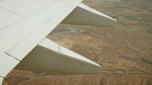 Ascending commercial airplane above arid countryside with wing and flap track fairings in foreground. Dry terrain and farmland below. Passenger flight gaining altitude after takeoff