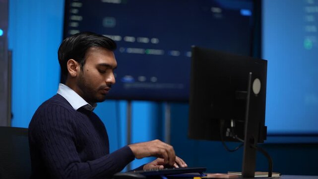 A developer reaching to his monitor adjusts something on screen and settles back to the keyboard in a blue lit room inside a dimly lit machine learning startup office.