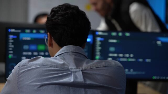 A developer seen from directly behind works through the dashboards while colleagues talk on out of focus behind him inside a dimly lit machine learning startup office.
