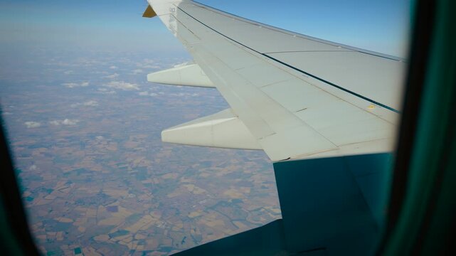 Passenger window view of airplane wing above patchwork farmland during high altitude flight. Rural landscape and scattered clouds below. Commercial aviation travel perspective