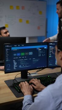 A seated developer typing in the foreground keeps working while a colleague gestures at the flowchart wall further back in the room at an AI company in Bangkok, Thailand.