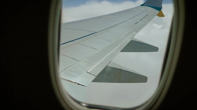Bright aircraft wing through passenger cabin window above dense white cloud layer. Airplane cruising in daylight over clouds. Peaceful air travel and aviation journey perspective