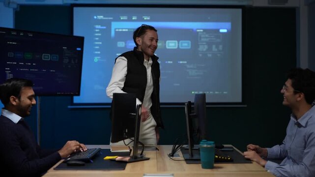 A team of three at their desks laugh together after a long run finally clears, monitors glowing across the frame inside a dimly lit machine learning startup office.