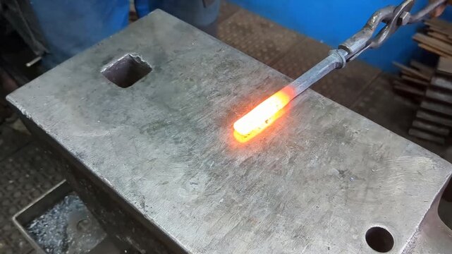A skilled metalworker uses traditional blacksmithing techniques to shape a glowing hot iron rod on a heavy steel anvil using a hammer and tongs in an industrial workshop environment