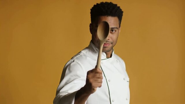 Camera rolling chef in jacket bringing wooden spoon to cover eye posing in mustard studio
