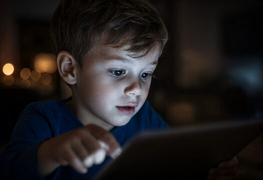 Young Boy Engaged With A Tablet, Eyes Lit Up In Amazement In A Dimly Lit Room.