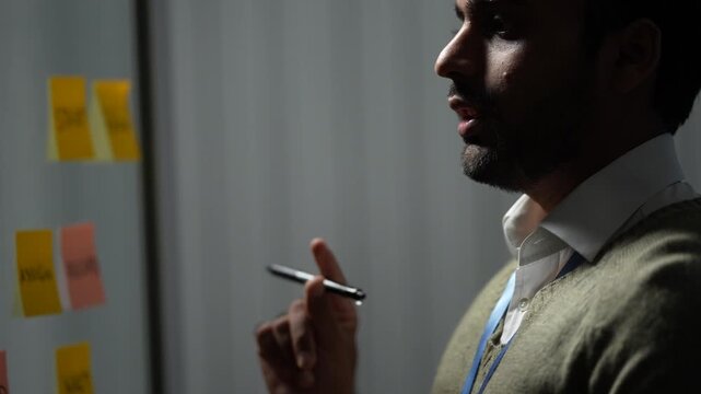 A bearded engineer in an olive sweater turns to the glass planning wall with a pen raised toward the sticky notes inside a dimly lit machine learning startup office.