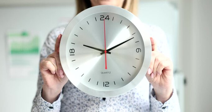 A person holds a large clock displaying the time, focusing on its numbers and hands. In a bright indoor setting, the passing minute signifies urgency and daily tasks ahead.
