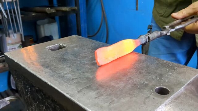 A skilled metalworker uses traditional blacksmithing techniques to shape a glowing hot iron rod on a heavy steel anvil using a hammer and tongs in an industrial workshop environment
