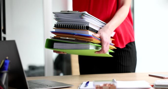In a bustling office, a person in a red top carefully balances a heavy stack of paperwork and notebooks. The sun shines through the windows, illuminating the workspace.