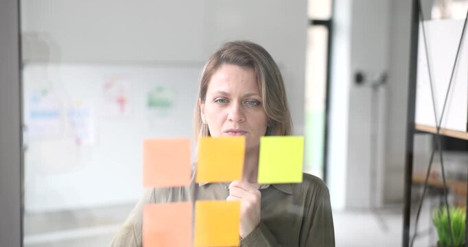 In a bright, airy office, a woman carefully organizes vibrant sticky notes on a glass panel. She looks focused and contemplative, capturing ideas and inspiration for her project.