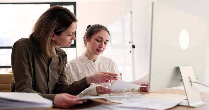 Two women engage in a lively discussion around a computer in a bright, contemporary office. They share ideas and review documents, fostering collaboration and innovation.