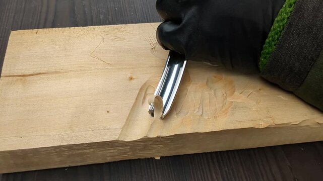 A gloved hand uses a curved metal chisel to carve intricate grooves into a flat wooden plank, creating small chips while shaping the material in a workshop environment with precision tools