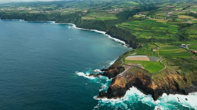 Wide aerial view of Sao Miguel coastline with Atlantic waves, volcanic cliffs and farmland near small lighthouse. Agricultural plots cover coastal terrain above ocean