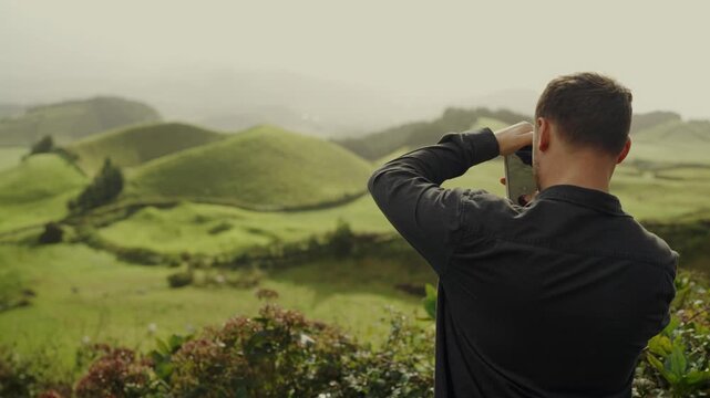 A rear view of a young man using binoculars to admire the scenic, verdant hills of Portugal, capturing the essence of exploration and nature. Man Using Binoculars to View Lush Green Hills in Portugal