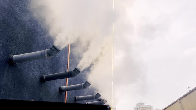 Industrial ventilation pipes expelling hot white steam or smoke from a building wall, contributing to air pollution and global warming under an overcast sky on a cold winter day