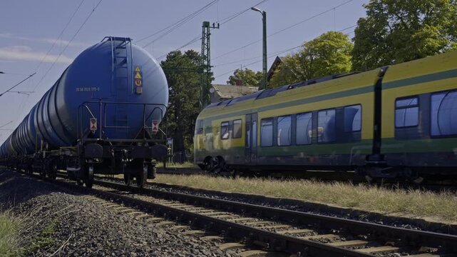 Blue railway tanker wagons and a passenger train travel on parallel tracks beneath overhead electric lines in a sunny rail transport scene.