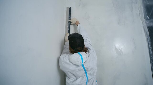 Professional construction worker wearing protective suit and gloves diligently applying plaster to wall with trowel, leveling surface during home renovation or repair project. 