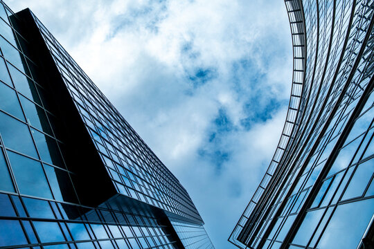 Modern skyscrapers viewed upward between glass facades where blue sky reflections and sharp architecture create dramatic geometric lines across the frame