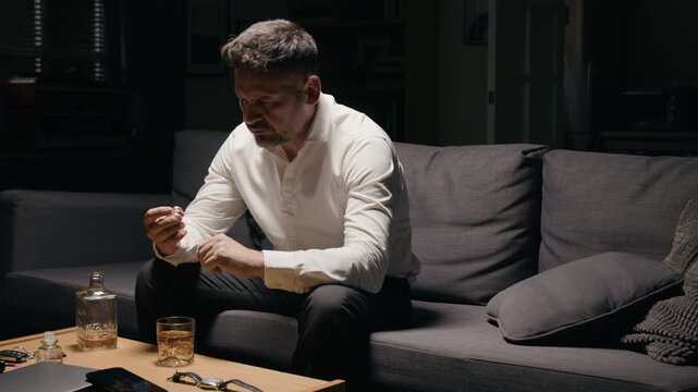 Middle aged Caucasian man examining wedding ring and drinking whiskey in distress. Footage suitable for content about divorce, alcohol addiction and mental health therapy campaigns
