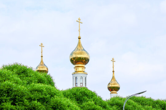Three ornate golden onion domes with crosses rise above lush green foliage against a pale sky; traditional Russian Orthodox architecture conveys spiritual grandeur and cultural heritage