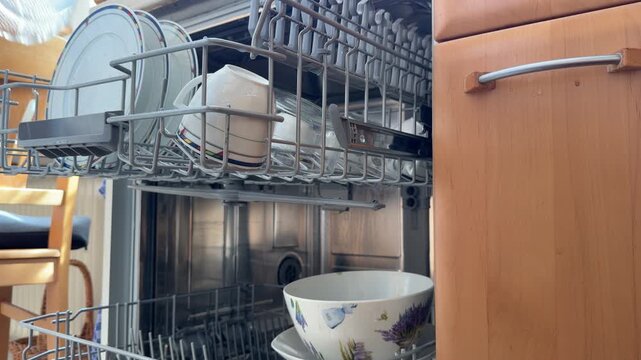 Low-angle close-up static shot of a woman's hand pulling out dishwasher racks and loading plates, cups, a bowl and a large dish into the open appliance at home.
