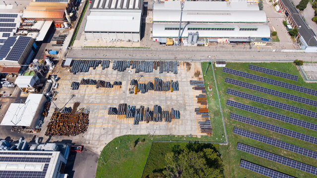 Aerial view of a metal construction facility with rows of steel pipes and industrial materials stored in an open yard, next to a solar panel array in Cornaredo, Lombardy, Italy.