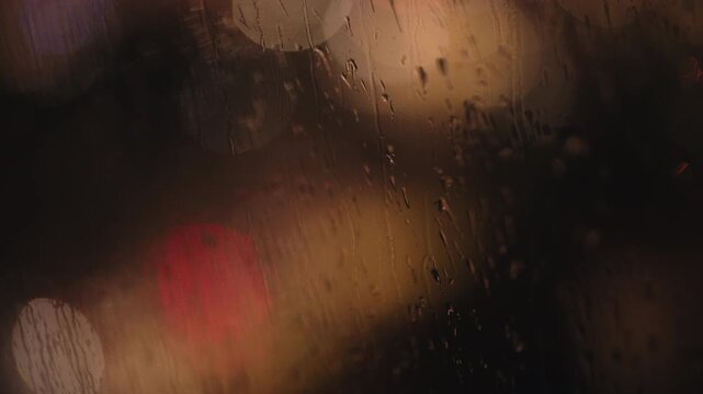 Raindrops dripping down a window pane during a dark, rainy night, creating a melancholic and peaceful mood with blurred city lights in the background creating a beautiful abstract bokeh effect