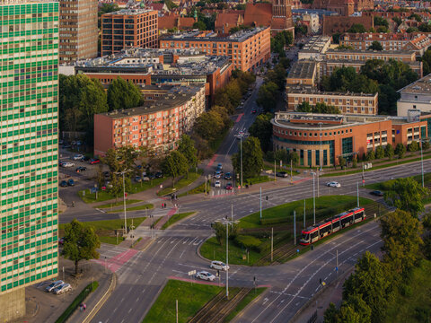 Aerial view of a busy urban intersection featuring a modern tram, a tall green skyscraper, and surrounding residential buildings in Gdansk, Pomeranian Voivodeship, Poland.