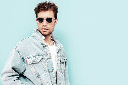 Portrait of handsome confident stylish hipster lambersexual model with curly hairstyle. Sexy man dressed in jeans jacket. Fashion male isolated on blue wall in studio. In sunglasses