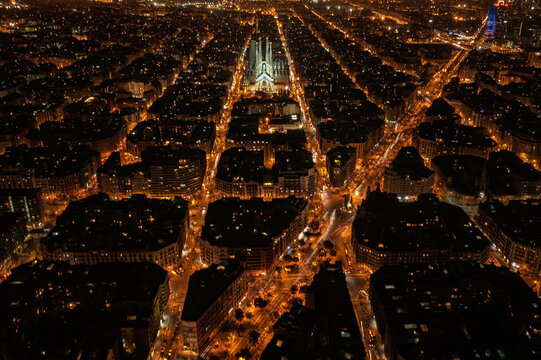 Aerial view of Sagrada Familia illuminated at night, showcasing the grid-like urban layout and glowing street lights of the Eixample district in Barcelona, Catalonia, Spain.
