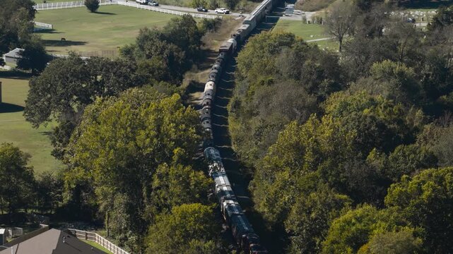 Long freight train traveling in wooded outskirts of Gentry, Arkansas, USA, aerial overview as caboose and tree shadows speckled on landscape