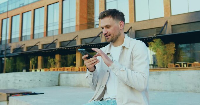 Close-up of a smiling young man in eyes glasses standing on a city street talking on the phone through a loudspeaker, recording a voice message against modern glass building outdoors