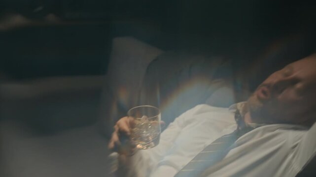 Caucasian middle aged businessman drinking whiskey from glass and falling asleep on couch. Footage suitable for mental health campaigns, addiction recovery programs, and workplace burnout awareness