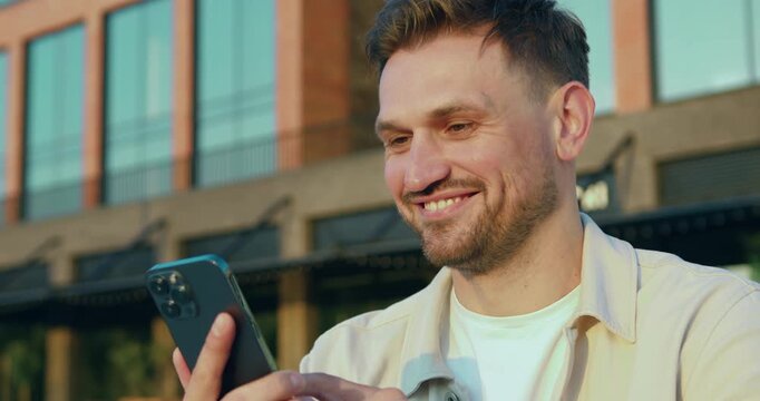 Handsome bearded Caucasian man using smartphone walking along city street near glass office building, outdoors