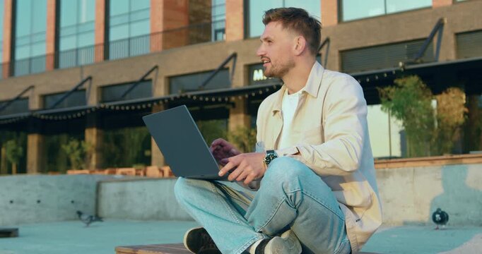 Successful Caucasian young man freelancer using laptop, watching webinars, working remotely, e-learning e-commerce online against in modern glass building background outdoors. Slow motion