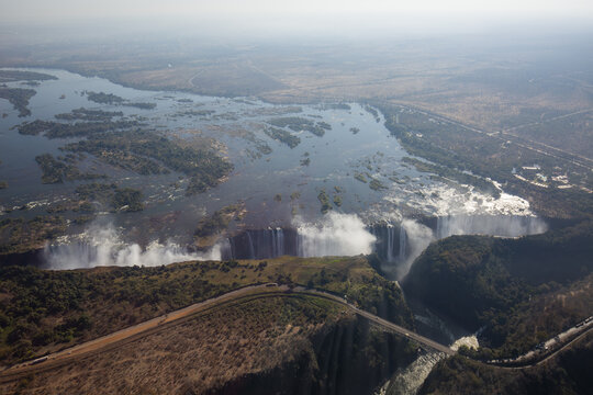 Aerial view of Victoria Falls with the Zambezi River flowing into the massive gorge, surrounded by lush vegetation and dry savanna landscape, Victoria Falls, Matabeleland North Pr, Zimbabwe.