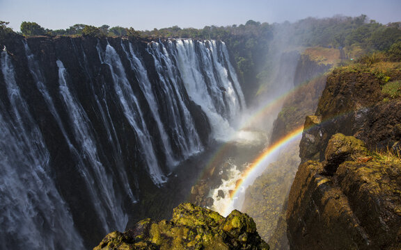 Aerial view of Victoria Falls cascading over rocky cliffs with a vibrant rainbow in the mist and lush vegetation in Victoria Falls, Matabeleland North Province, Zimbabwe.