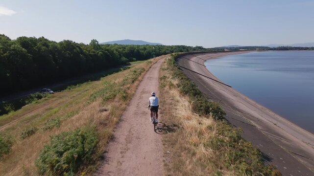 Cyclist riding bicycle on gravel road beside lake, aerial view