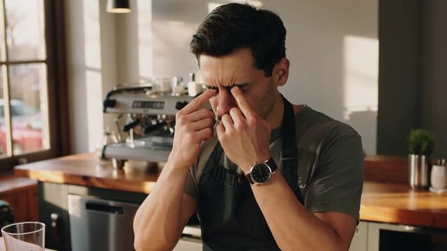 Warm sunlight from window, barista rubbing eyes and pressing brows at cafe counter